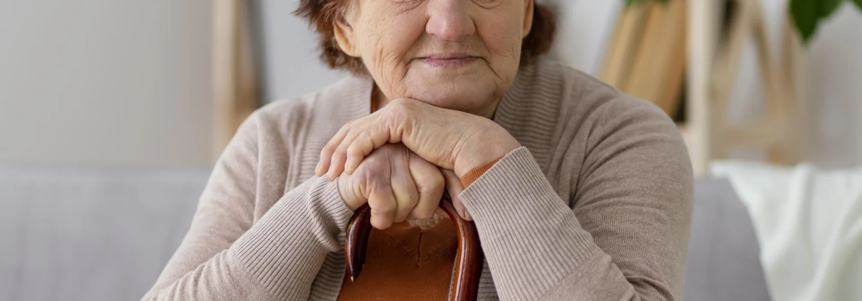 Une femme senior assise chez elle, les mains posées sur sa canne.