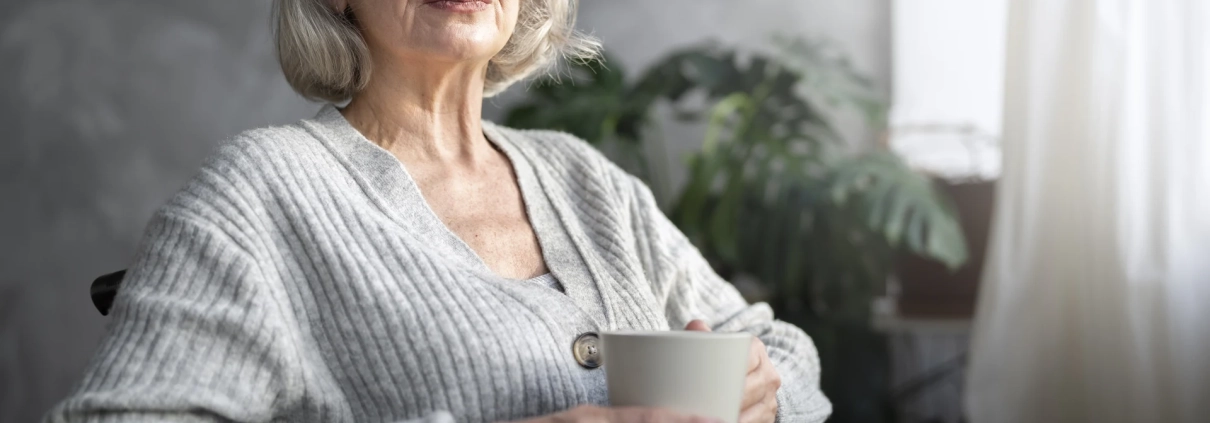 Une femme senior assise dans un fauteuil roulant tient une tasse près d’une fenêtre.