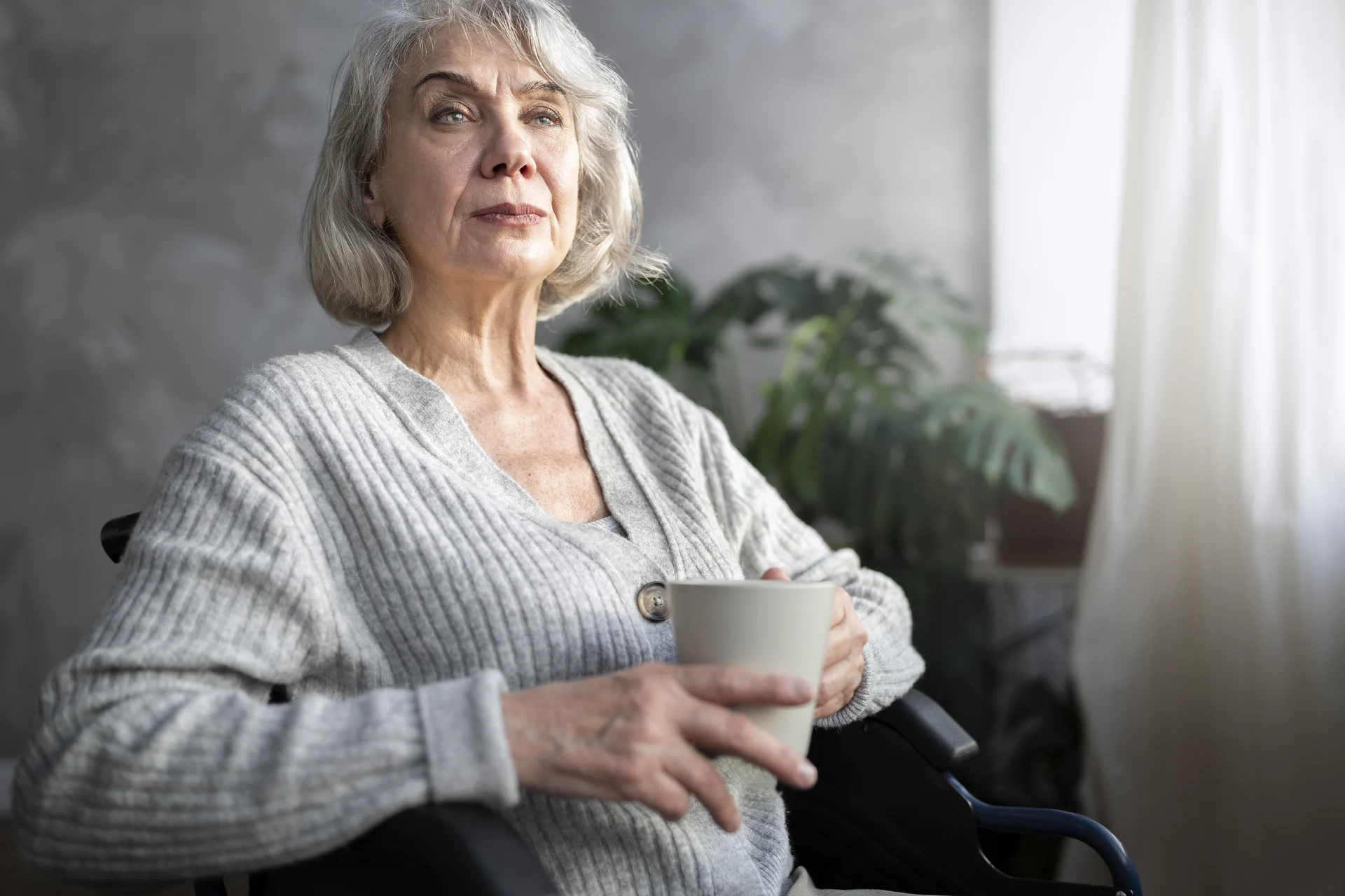 Une femme senior assise dans un fauteuil roulant tient une tasse près d’une fenêtre.